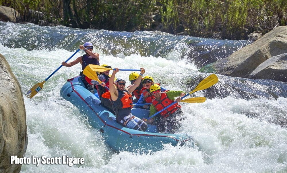 Whitewater rafting guide pilots a family of five excited adults and kids clutching paddles through churning rapids in a bright blue raft